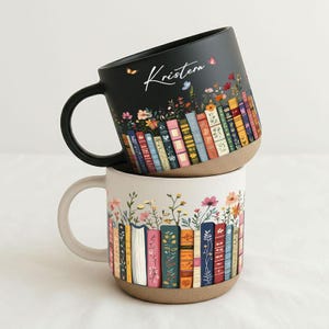 Könnte beinhalten: Zwei Keramikbecher, einer schwarz und einer cremefarben, gestapelt. Der schwarze Becher hat den Namen "Kristen" in Schreibschrift und ein Blumen- und Buchdesign. Der cremefarbene Becher hat ein ähnliches Design. Beide Becher haben einen braunen Boden und einen gebogenen Griff.