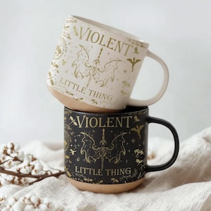 Könnte beinhalten: Zwei gestapelte Keramikbecher. Der obere Becher ist cremefarben mit goldfarbenen Buchstaben und einer fledermausartigen Grafik mit der Aufschrift "VIOLENT LITTLE THING". Der untere Becher ist schwarz mit dem gleichen Design. Beide Becher haben einen beigefarbenen Boden und einen Henkel.
