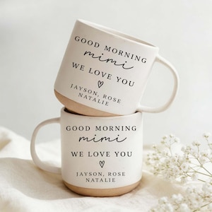 Könnte beinhalten: Zwei gestapelte weiße Keramikbecher mit beigem Boden. Jeder Becher trägt den Text "GOOD MORNING mimi WE LOVE YOU ♡ JAYSON, ROSE NATALIE" in schwarzer Schrift. Die Becher haben ein schlichtes, elegantes Design.