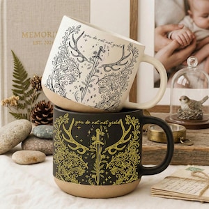 May include: Two ceramic mugs, one white and one black, stacked. The white mug features a black antler and sword design with the text "you do not not yield." The black mug has a gold antler and sword design with the same text. Both mugs have a tan base.