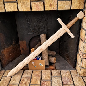 Op de afbeelding: Een houten speelgoedzwaard met een lichtgekleurd lemmet met een centrale richel, een rechte pareerstang en een handvat omwikkeld met natuurlijk bruin touw. De pommel is een eenvoudig, plat houten blok, vervaardigd uit glad, onafgewerkt licht hout.