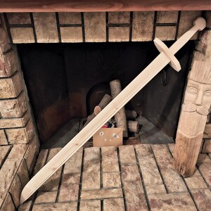 Puede incluir: Una espada de juguete de madera de color claro que presenta una hoja larga y recta, una guarda transversal sencilla y un pomo redondeado. La espada está fabricada en madera natural con vetas visibles, mostrando un acabado liso y pulido en toda la pieza.