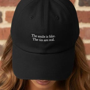 May include: Black baseball cap with the text "The smile is fake. The tits are real." in white. The cap is worn by a person with long, wavy brown hair, against a brick wall background.