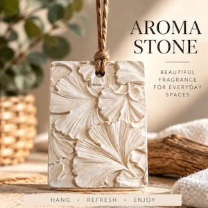 May include: A rectangular off-white aroma stone featuring a relief pattern of ginkgo leaves. The stone hangs from a brown jute twine cord. Text on the image reads AROMA STONE, BEAUTIFUL FRAGRANCE FOR EVERYDAY SPACES, and HANG • REFRESH • ENJOY.