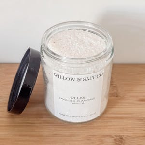 Peut inclure: Un pot en verre transparent rempli de sels de bain minéraux blancs. Le pot a un couvercle noir posé à côté. L'étiquette indique "Willow & Salt Co. RELAX Lavande, Camomille, Vanille Bain Minéral 227g."