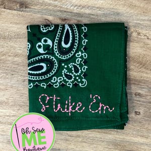 May include: A dark green bandana featuring a white paisley pattern and the text Strike 'Em embroidered in pink thread. The fabric is folded into a square shape, displaying the intricate paisley design in the corner and the stitched lettering.
