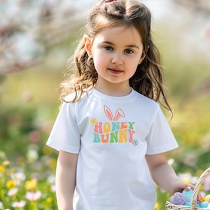 May include: A young girl wearing a white t-shirt with the words "HONEY BUNNY" in colorful, retro-style lettering. The shirt features bunny ears and flower graphics. The girl is holding a basket with Easter eggs.