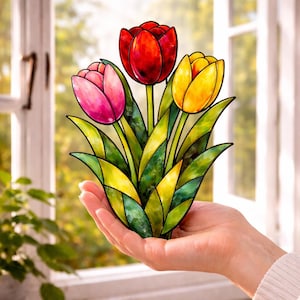 Puede incluir: Un ramo de tulipanes de vidriera con tres flores rojas, rosas y amarillas. Las hojas son verdes y amarillas. La obra de arte se sostiene en una mano, con una ventana y vegetación en el fondo.