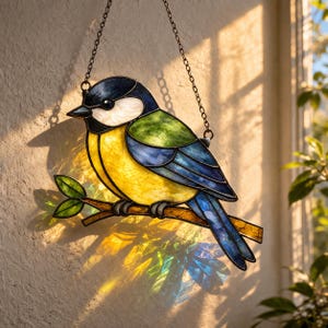 Könnte beinhalten: Buntglas-Vogelsonnenfänger. Der Vogel hat eine gelbe Brust, blaue Flügel und einen schwarzen Kopf. Er sitzt auf einem braunen Ast mit grünen Blättern. Der Sonnenfänger hängt an einer Metallkette und wirft farbiges Licht.