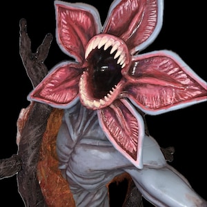 May include: A creature figurine featuring a large, open, flower-like head with five pink, fleshy petals and sharp, white, pointed teeth surrounding a dark, hollow center. The body is a pale, grayish-blue tone with a textured, dark brown, root-like structure.