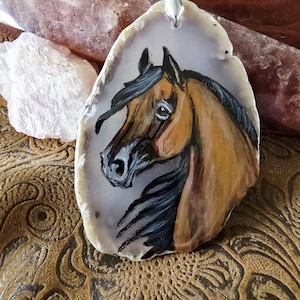 Op de afbeelding: Handbeschilderde paardenhanger op een natuurlijke agaatsteen. Het paard is afgebeeld in bruin- en zwarttinten, met een zilveren beugel aan de bovenkant. De steen heeft een lichtgrijs en wit patroon. De hanger is een uniek sieraad.