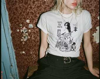 May include: A white short-sleeve t-shirt featuring a black graphic print of a figure with a snake and the text 'both the one's' and 'in the nameless sodality'. The shirt is paired with black trousers featuring a black belt with a gold-toned buckle.