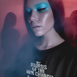 Puede incluir: Una persona con una camiseta negra con el texto "THE LORDS OF THE NEW CHURCH" en blanco. La persona tiene el pelo largo y negro y sombra de ojos azul brillante. El fondo está borroso con tonos rojos y azules.