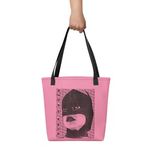 Pu&ograve; includere: Una borsa tote rosa con manici neri caratterizzata da una stampa grafica nera di una persona che indossa un cappuccio e una maschera scuri. Il testo NINA HAGEN &egrave; stampato verticalmente in nero lungo il lato sinistro del grafico su un motivo.