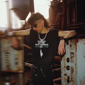 Puede incluir: Camiseta negra con el texto "LORDS OF THE NEW CHURCH" y un gráfico de espada. La persona lleva gafas de sol y un collar de cadena plateada. El fondo es un entorno industrial.