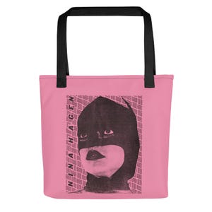 Pu&ograve; includere: Una borsa tote rosa con manici neri caratterizzata da una stampa grafica nera di una persona che indossa un cappuccio scuro e il testo NINA HAGEN stampato verticalmente sul lato sinistro. Lo sfondo del grafico consiste in un motivo a linee orizzontali.