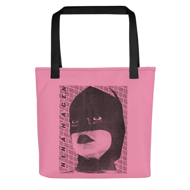 Pu&ograve; includere: Una borsa tote rosa con manici neri caratterizzata da una stampa grafica nera di una persona che indossa un cappuccio scuro e il testo NINA HAGEN stampato verticalmente sul lato sinistro. Lo sfondo del grafico consiste in un motivo a linee orizzontali.
