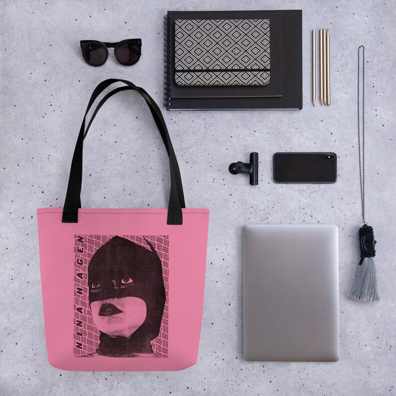 Pu&ograve; includere: Una borsa tote rosa con manici neri caratterizzata da una stampa grafica nera di una persona che indossa una maschera e il testo Nina Hagen. La borsa &egrave; mostrata insieme a occhiali da sole, un taccuino, uno smartphone, un portatile e una lunga collana con nappa.