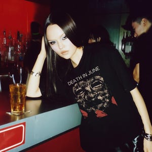 Può includere: Una t-shirt nera a maniche corte con la scritta bianca DEATH IN JUNE sopra la grafica di un volto scuro e strutturato simile a una maschera, con accenti rossi sul fondo. Indossata con un braccialetto color argento al polso.