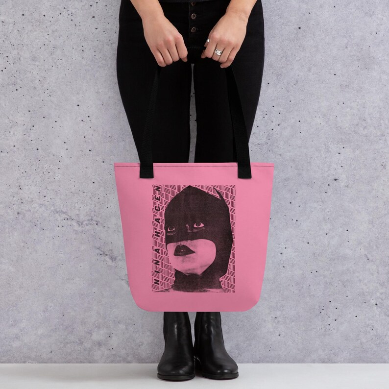 Pu&ograve; includere: Una borsa tote rosa con una stampa grafica nera di una persona che indossa un cappuccio scuro. Il design include il testo NINA HAGEN verticalmente sul lato sinistro del ritratto, su uno sfondo di motivi geometrici a linee ripetute.