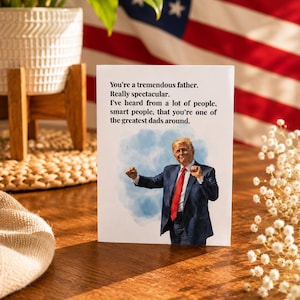 May include: A white greeting card featuring a watercolor-style illustration of a man in a navy blue suit and red tie with both fists raised. The text reads: You're a tremendous father. Really spectacular. I've heard from a lot of people.
