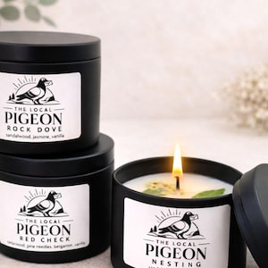 Op de afbeelding: Drie zwarte, ronde kaarsen met witte etiketten met een duivenlogo en geurbeschrijvingen. E&eacute;n kaars is aangestoken en toont een vlam en een wit wasinterieur. De etiketten luiden "Rock Dove", "Red Check" en "Nesting".
