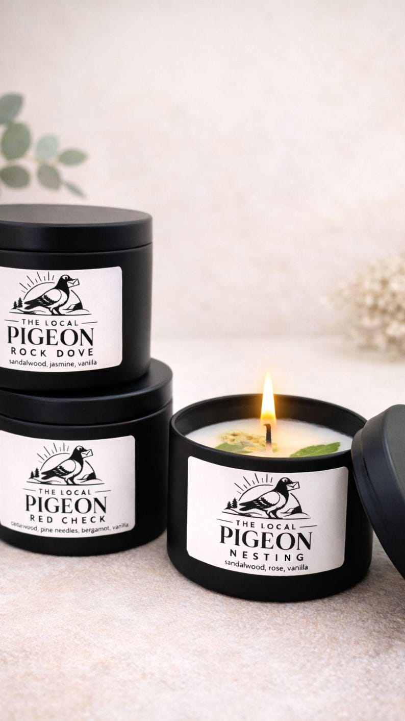 Op de afbeelding: Drie zwarte, ronde kaarsen met witte etiketten met een duivenlogo en geurbeschrijvingen. E&eacute;n kaars is aangestoken en toont een vlam en een wit wasinterieur. De etiketten luiden "Rock Dove", "Red Check" en "Nesting".