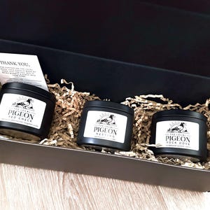 Op de afbeelding: Een zwarte geschenkdoos met drie zwarte kaarsblikken met witte etiketten. De etiketten lezen "The Local Pigeon" met verschillende geurnamen. De doos is gevuld met bruin papier en een kleine bedankkaart.