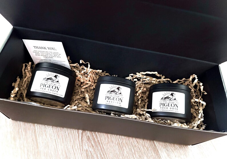 Op de afbeelding: Een zwarte geschenkdoos met drie zwarte kaarsblikken met witte etiketten. De etiketten lezen "The Local Pigeon" met verschillende geurnamen. De doos is gevuld met bruin papier en een kleine bedankkaart.
