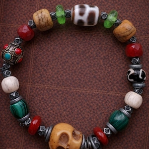 Puede incluir: Pulsera de cuentas con un dije de calavera y varias cuentas rojas, verdes, marrones y blancas. La pulsera presenta una mezcla de texturas y formas, con detalles metálicos plateados.