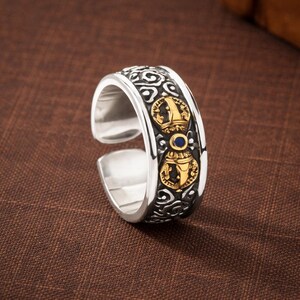 Puede incluir: Un anillo ajustable de plata con detalles ornamentados. El anillo presenta una banda central con detalles dorados y una pequeña piedra azul. El diseño incluye patrones y símbolos intrincados, creando una joya única y llamativa.