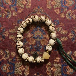 May include: A cream-colored skull bead bracelet with brass accents and a green cord. The bracelet features a small, heart-shaped yellow stone and is displayed on a patterned red and blue fabric.