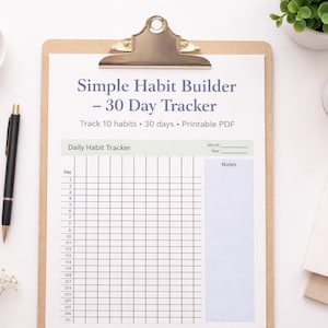 May include: A clipboard with a "Simple Habit Builder - 30 Day Tracker" printable PDF. The tracker includes space to track 10 habits over 30 days. A black pen, a small bowl of gold paper clips, and a small plant are also visible.