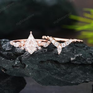 May include: Two rose gold rings. One ring has a kite-shaped, clear gemstone with smaller stones. The second ring has a curved design with clear gemstones. The rings are displayed on a dark, textured surface.