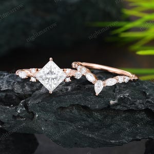 May include: A rose gold engagement ring set with a princess-cut centre stone and a curved band with smaller accent stones. The rings are displayed on a dark, textured surface.
