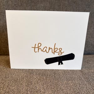 May include: A white greeting card featuring the word thanks in gold cursive lettering positioned above a black graphic illustration of a rolled diploma tied with a ribbon. The card has a clean, minimalist design with a smooth, matte finish.