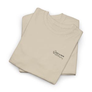 Könnte beinhalten: Zwei gefaltete, beigefarbene T-Shirts mit Rundhalsausschnitt und kurzen Ärmeln. Auf der Vorderseite der Shirts steht "JUST A MOM", zusammen mit einer Entengrafik und den Worten "KEEPING HER DUCKS IN A ROW".