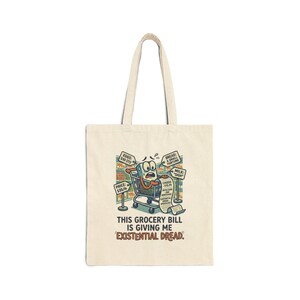 May include: Cream-colored tote bag with a cartoon grocery cart design. The cart has a worried expression, surrounded by signs listing grocery items and prices. Text on the bag reads, "THIS GROCERY BILL IS GIVING ME EXISTENTIAL DREAD."