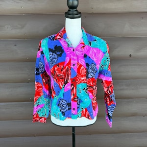 Pode incluir: Uma camisa abotoada com um padrão floral vibrante em vermelho, rosa, azul e verde-azulado. A camisa de manga comprida apresenta um colarinho e botões pretos. A estampa floral inclui rosas e outras flores estilizadas.