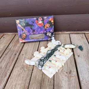 May include: A set of white plastic ghost-shaped novelty string lights with a dark green cord, packaged in a rectangular cardboard box featuring colorful illustrations of children in Halloween costumes and the text Halloween Novelty Lights.