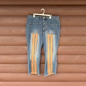 May include: Blue denim jeans with orange vertical panels down the front and back of the legs. The jeans have a classic five-pocket design, a button and zipper closure, and are hanging on a white hanger.