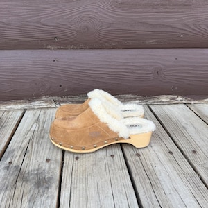 May include: A pair of brown suede clogs with a shearling lining and wooden soles. The clogs have a rounded toe and are adorned with small silver studs around the sole. The brand name "UGG" is visible on the side.