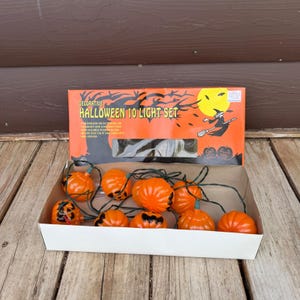 May include: A decorative Halloween light set in a cardboard box featuring ten orange pumpkin-shaped light covers. The box displays a yellow moon, a witch on a broomstick, and the text DECORATIVE HALLOWEEN 10 LIGHT SET with black tree silhouettes.