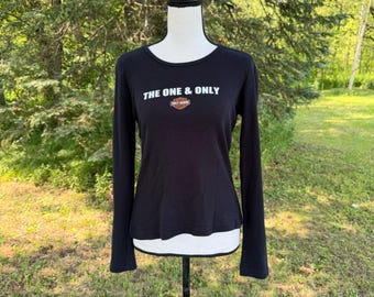 May include: A black long-sleeve shirt featuring a crew neckline and white text that reads THE ONE & ONLY above a small orange, white, and black Harley-Davidson logo graphic centered on the chest. The shirt is displayed on a white mannequin.