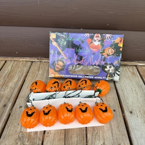 Puede incluir: Una cadena de diez cubiertas de luz de plástico naranja con forma de calabaza y rasgos faciales pintados en negro, conectados por un cable verde. Las luces están empaquetadas en una caja de cartón rectangular con ilustraciones de niños disfrazados y el texto Halloween Novelty Lights.