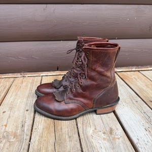 Könnte beinhalten: Ein Paar knöchelhohe Schnürstiefel aus braunem Used-Look-Leder. Die Stiefel haben eine abgerundete Zehenpartie, einen geschichteten Holzabsatz, dunkle Metallösen mit passenden braunen Schnürsenkeln und eine dekorative Lederfransenlasche.