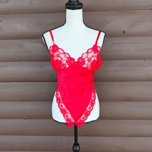 May include: A red lingerie bodysuit with delicate lace detailing. The bodysuit features thin straps and lace trim along the neckline and leg openings. The garment is displayed on a white mannequin against a brown wooden background.