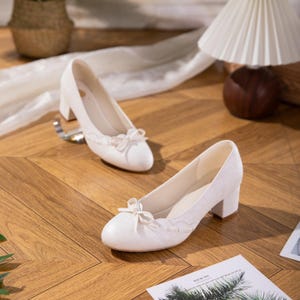 Könnte beinhalten: Ein Paar elfenbeinfarbene Pumps mit Blockabsatz. Jeder Schuh hat eine kleine Schleife an der Spitze und dekorative Verzierungen am oberen Rand. Die Schuhe sind aus einem strukturierten Material und stehen auf einem Holzboden.