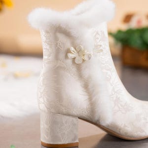 Peut inclure: Une bottine blanc cass&eacute; avec un motif floral ton sur ton subtil, une bordure &eacute;paisse en fausse fourrure blanche le long du col et de la couture lat&eacute;rale, et une fermeture d&eacute;corative en tissu en forme de fleur. Talon bloc et finition lisse.