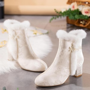 Peut inclure: Une paire de bottines couleur cr&egrave;me avec un motif floral jacquard subtil, une bordure en fausse fourrure blanche autour de l'ouverture sup&eacute;rieure et une fleur d&eacute;corative sur le c&ocirc;t&eacute;. Les bottines ont un talon bloc et un bout arrondi.